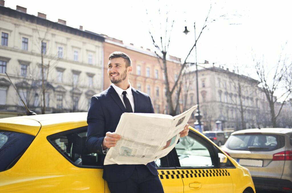 pexels photo 1843360 A businessman in a suit reads a newspaper while leaning on a yellow taxi in a city street.
